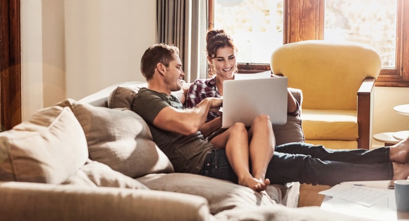 Ein junges Paar sitzt auf seiner Couch und informiert sich per Laptop im Internet über Eigentumswohnungen als Wertanlage.